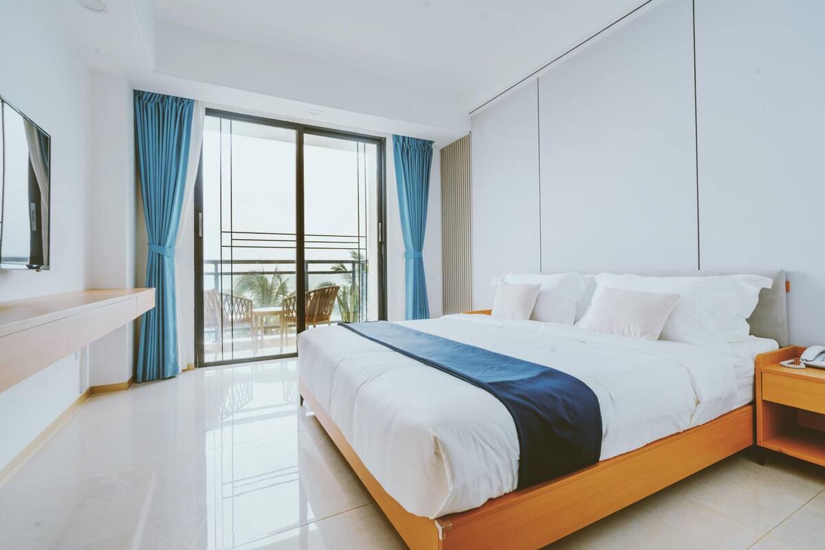 A modern hotel room with a large bed, a bedside table, a mirror, and a balcony with curtains.