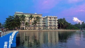 A multi-story hotel with balconies, located beside a calm body of water, surrounded by palm trees and a clear sky. - Angel Garden Resort Coron (Busuanga)