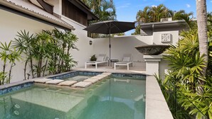 A swimming pool with a stone border, surrounded by palm trees and a lounge area with chairs and an umbrella.