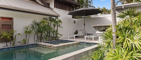 A swimming pool with a tiled deck, surrounded by tropical plants and a white building with a sloped roof.