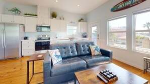 A modern kitchen with white cabinets, a stainless steel refrigerator, and a microwave. There is a blue leather sofa with decorative pillows, a wooden coffee table, and a sign above the windows that reads "Welcome to Jay's Surf Shack." - Shared Pool, Golf Cart Access, Pet Friendly (Port Aransas)
