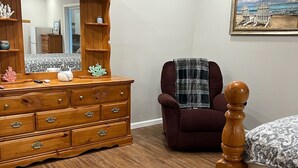 Een slaapkamer met een houten dressoir, een stoel, een bed, een spiegel en een schilderij aan de muur.