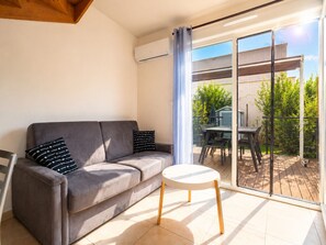 A modern living room with a grey sofa, a small round table, and a sliding glass door leading to an outdoor patio with a table and chairs.