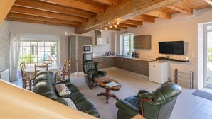 A spacious living area with a wooden ceiling, a sofa set, a dining table with chairs, a kitchen with modern appliances, and a TV mounted on the wall.