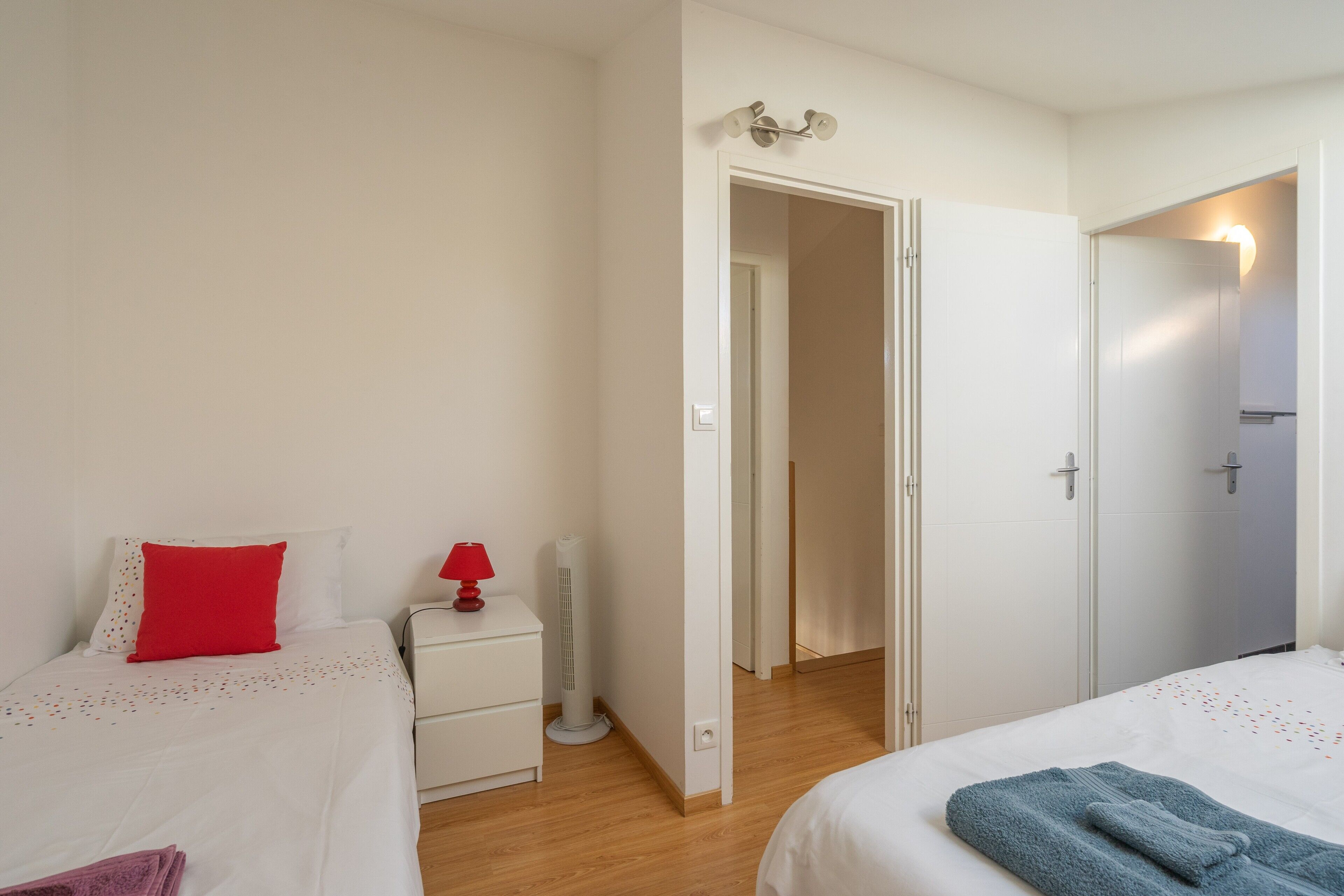 Un dormitorio con dos camas, una mesita de noche con lámpara, un calentador y una puerta que da a otra habitación.