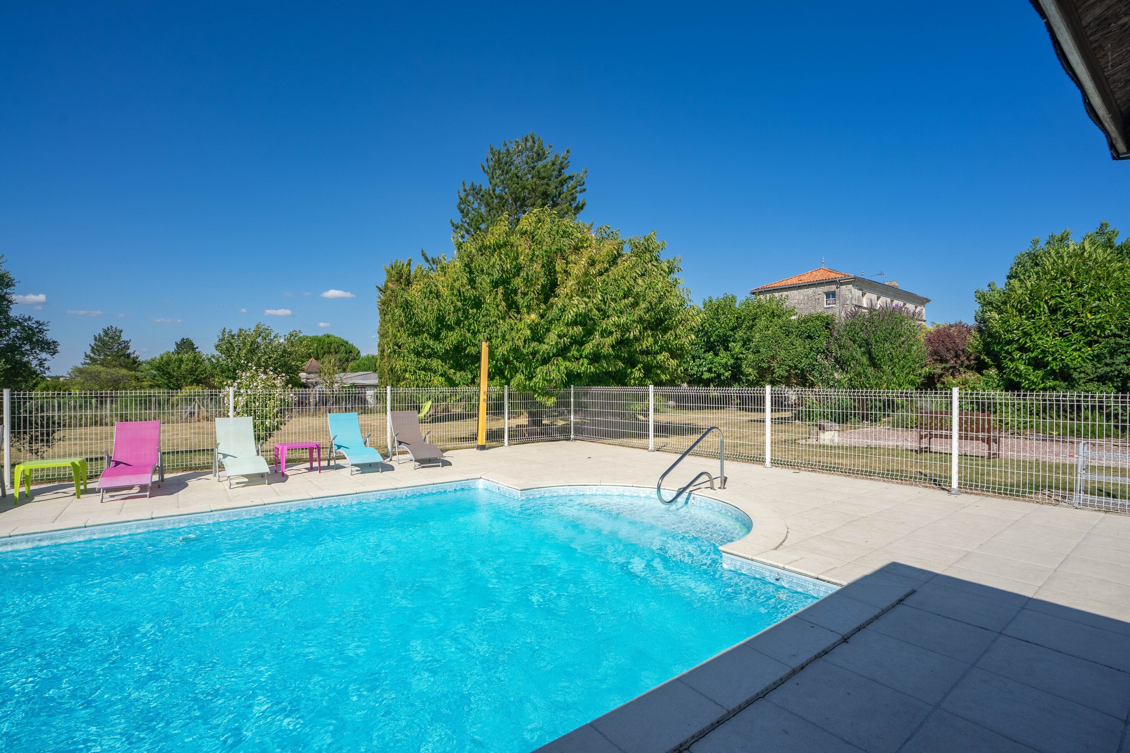 A swimming pool with a fenced deck, surrounded by trees and a building in the background.