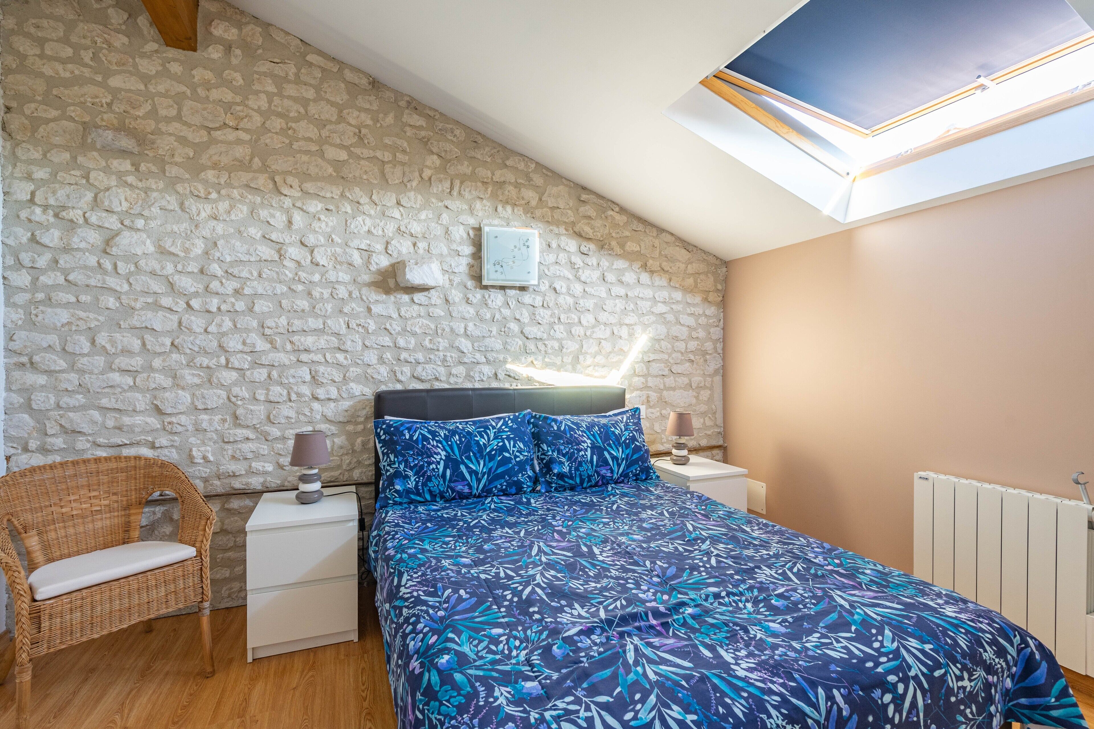 Una camera da letto con parete in pietra, un letto con copripiumino floreale, una sedia in vimini, un comodino con una lampada e un lucernario.