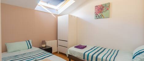 A bedroom with two beds, a skylight, a radiator, a wardrobe, and a painting on the wall.