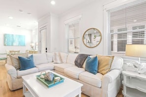 A modern living room with a white sectional sofa, a coffee table, and a dining area in the background.
