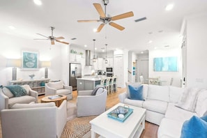 A modern living room with white furniture, a kitchen in the background, and ceiling fans.