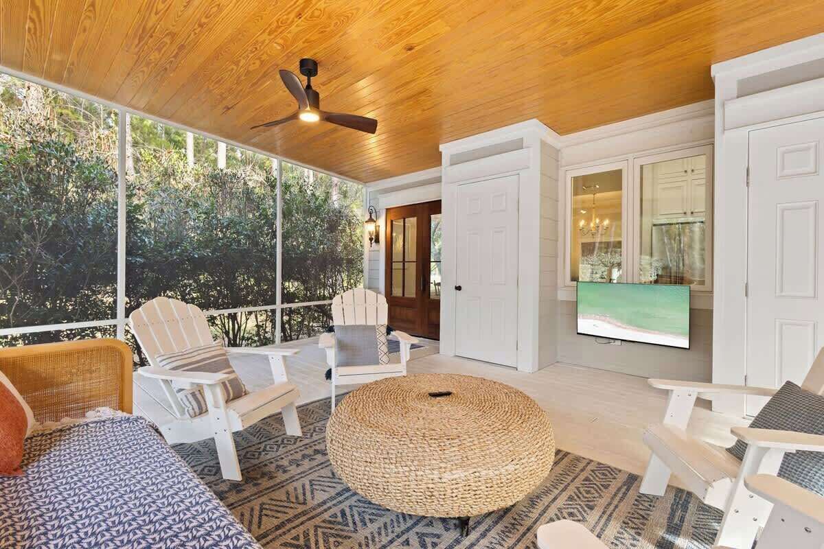 A covered outdoor seating area with wicker furniture, a round ottoman, and a television screen.