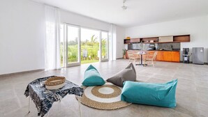 Un vaste espace de vie avec une grande porte-fenêtre coulissante donnant sur un balcon, une cuisine équipée d’appareils électroménagers modernes et un coin repas avec des chaises en osier.