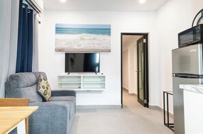 Una sala de estar moderna con un sofá gris, un televisor de pantalla plana montado en la pared y una cocina con microondas y refrigerador.