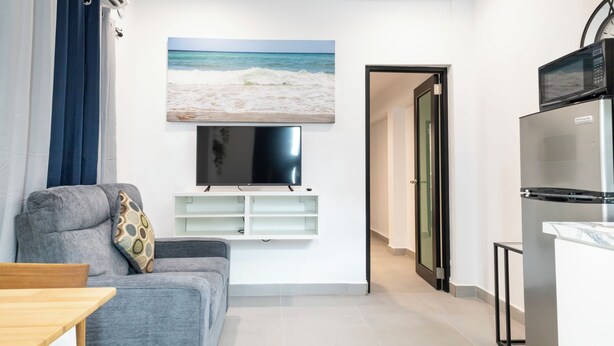 Una sala de estar moderna con un sofá gris, un televisor de pantalla plana montado en la pared y una zona de cocina con refrigerador y microondas.