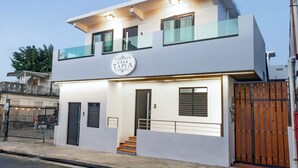 Un edificio moderno de dos pisos con un cartel que dice "Casa Tapia". Cuenta con un balcón, una entrada con portón y una escalera que lleva a la entrada.