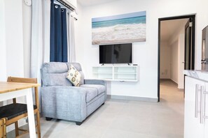 Una sala de estar con un sofá gris, una mesa de comedor de madera, un televisor de pantalla plana montado en la pared y una zona de cocina con armarios blancos.