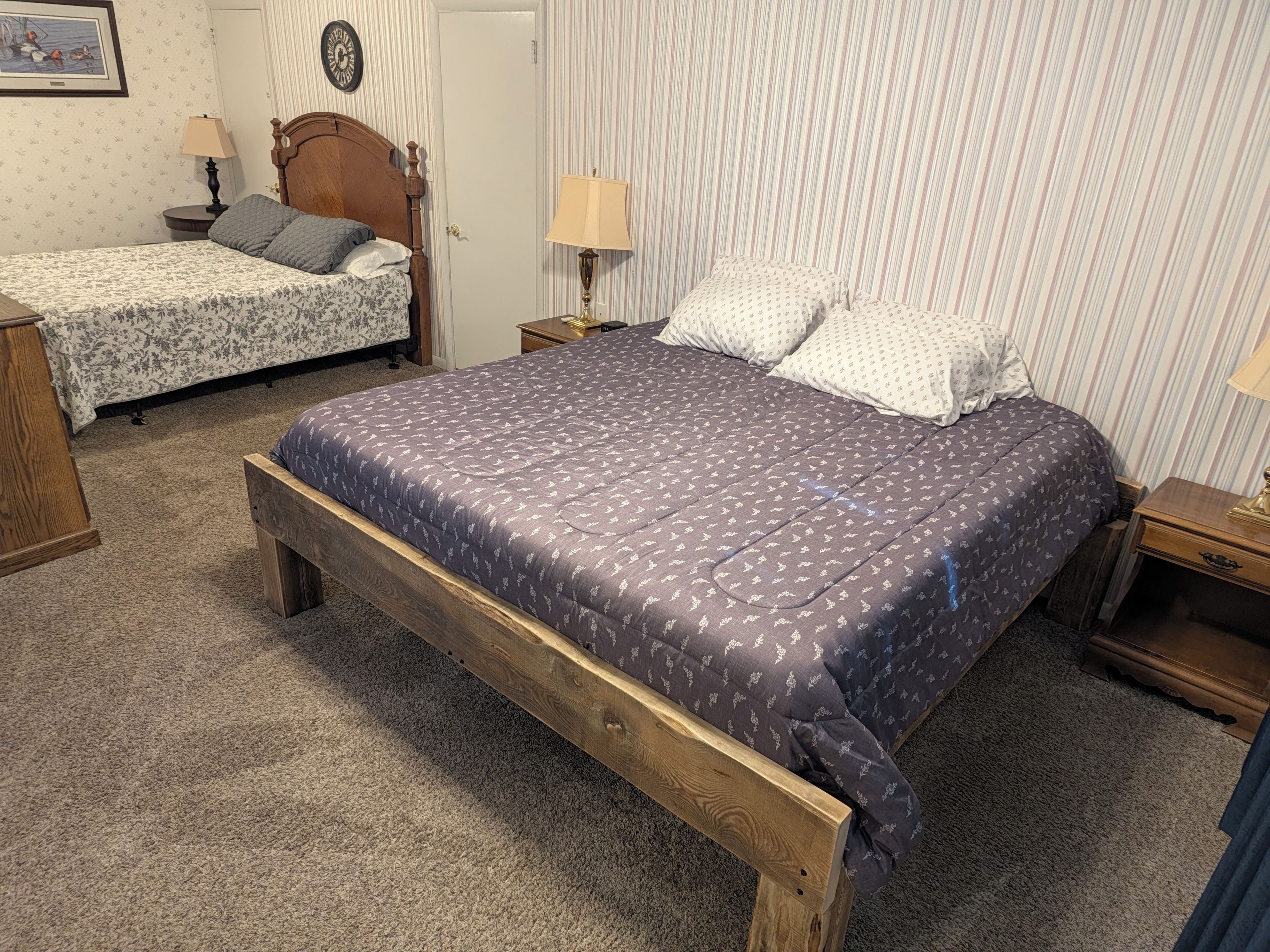 Une chambre à coucher avec un grand lit, un plus petit lit, deux tables de chevet, une commode, une lampe, une horloge et un tableau encadré au mur.