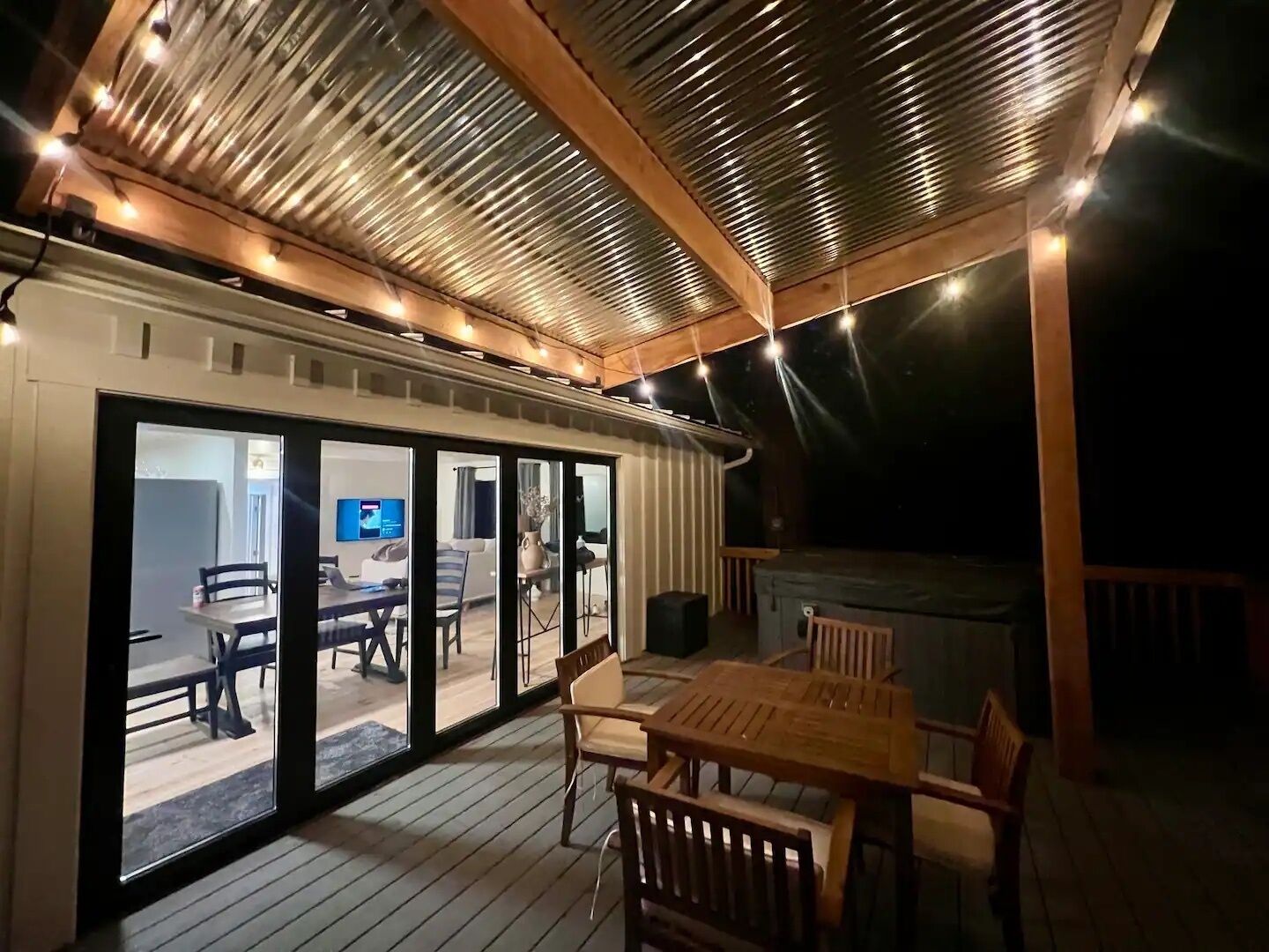 A covered outdoor area with a wooden deck, a hot tub, and a dining table with chairs.