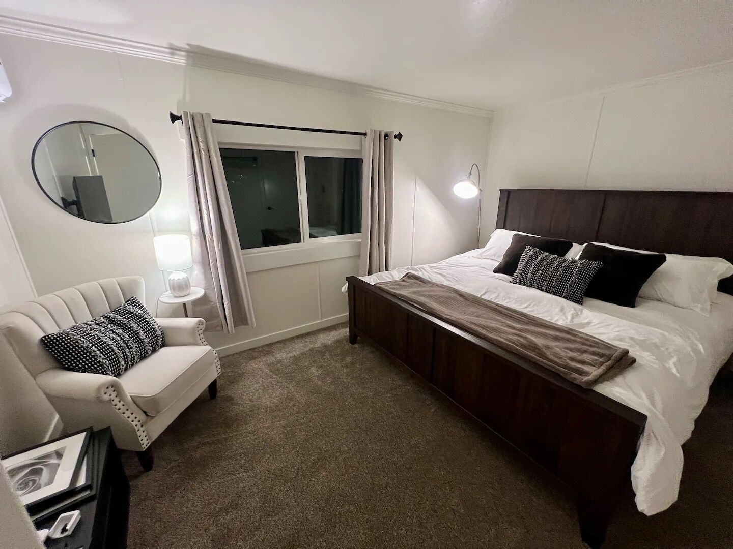 A bedroom with a bed, a chair, a bedside table, a lamp, a window with curtains, and a mirror.
