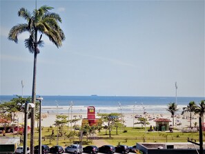 A beach view with palm trees, a red building, and parked cars. - Holiday Flat in São Vicente (Itararé São Vicente)