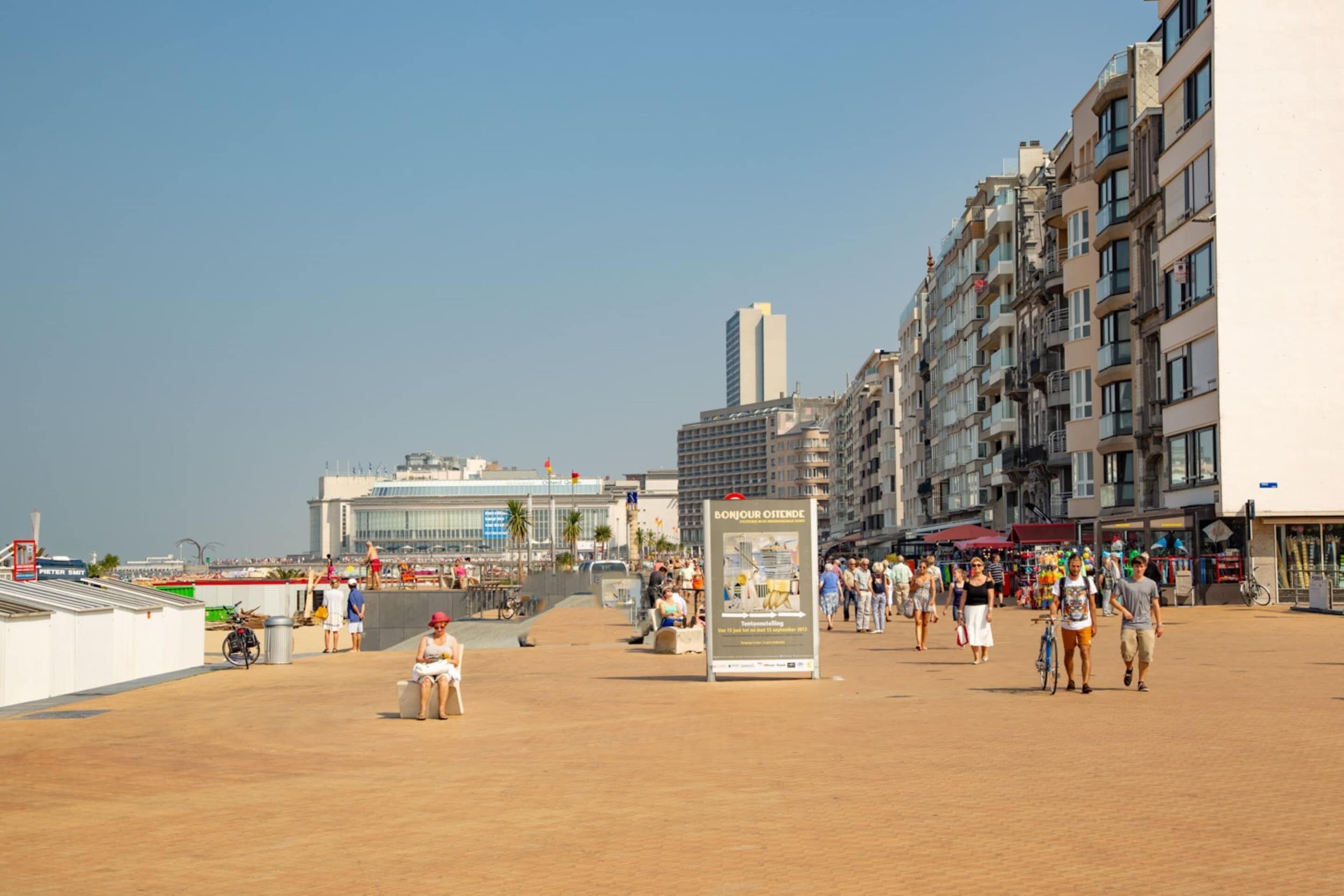 A sandy beach with people walking, a signboard, and buildings in the background.