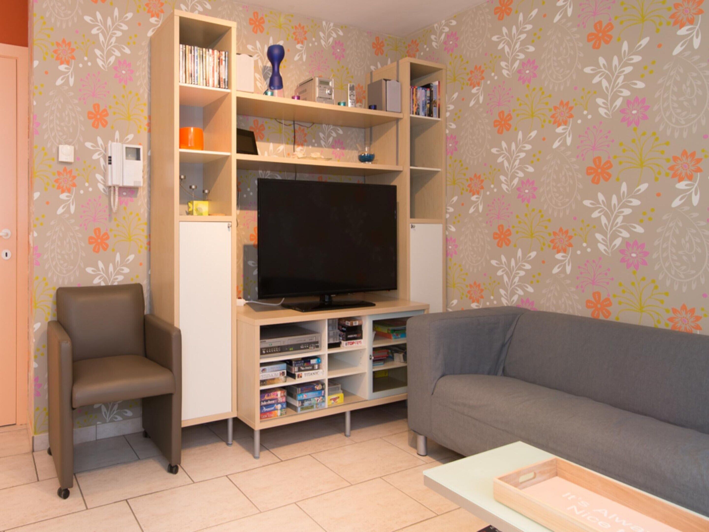 A living room with a flat-screen TV, a grey sofa, a brown chair, and a bookshelf with various items.