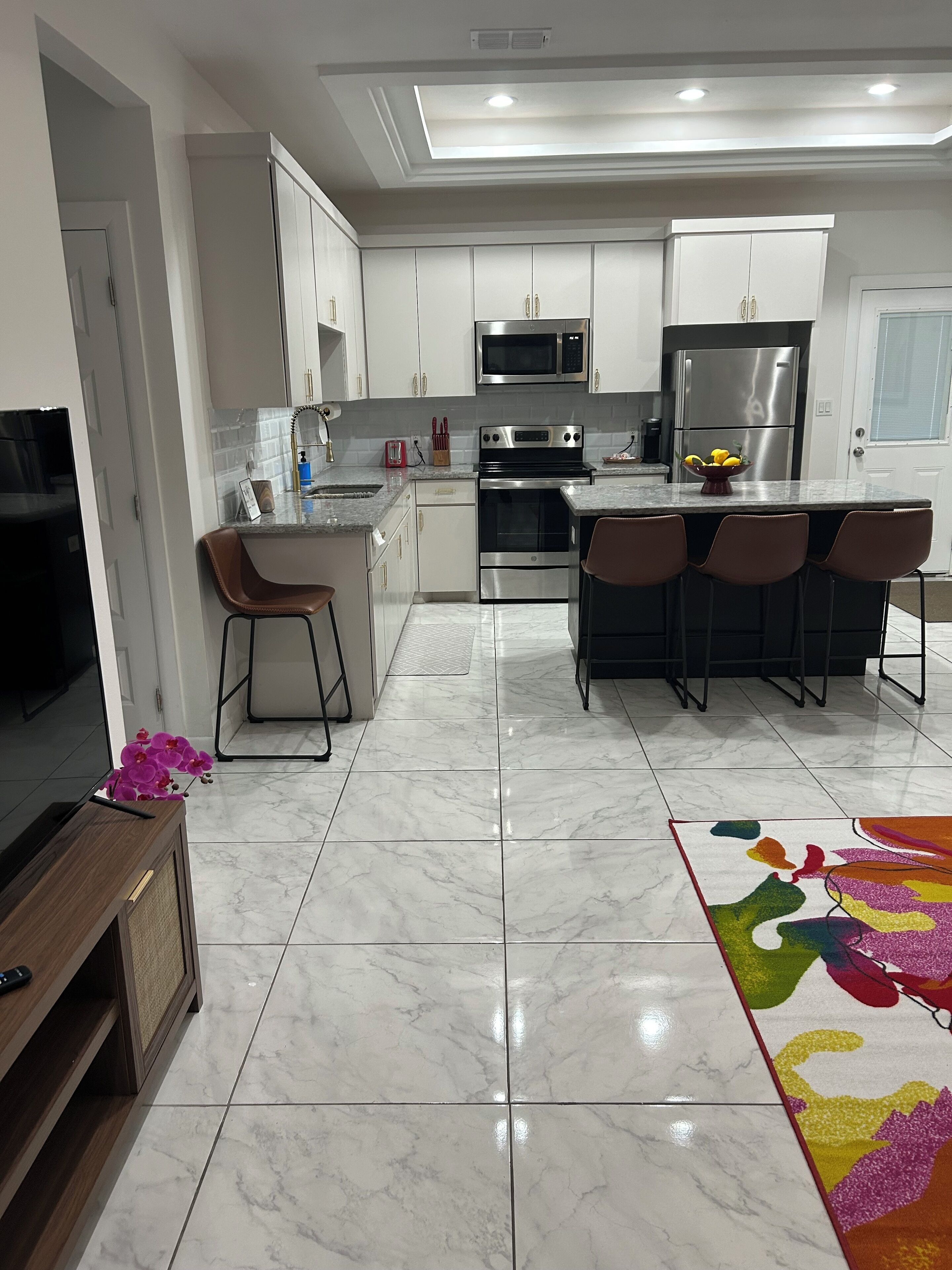 Una cocina moderna con armarios blancos, electrodomésticos de acero inoxidable, piso de mármol y una alfombra colorida.