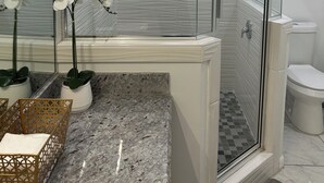 Un baño moderno con ducha cubierta por vidrio, azulejos blancos, encimera de mármol y sanitarios en color blanco.