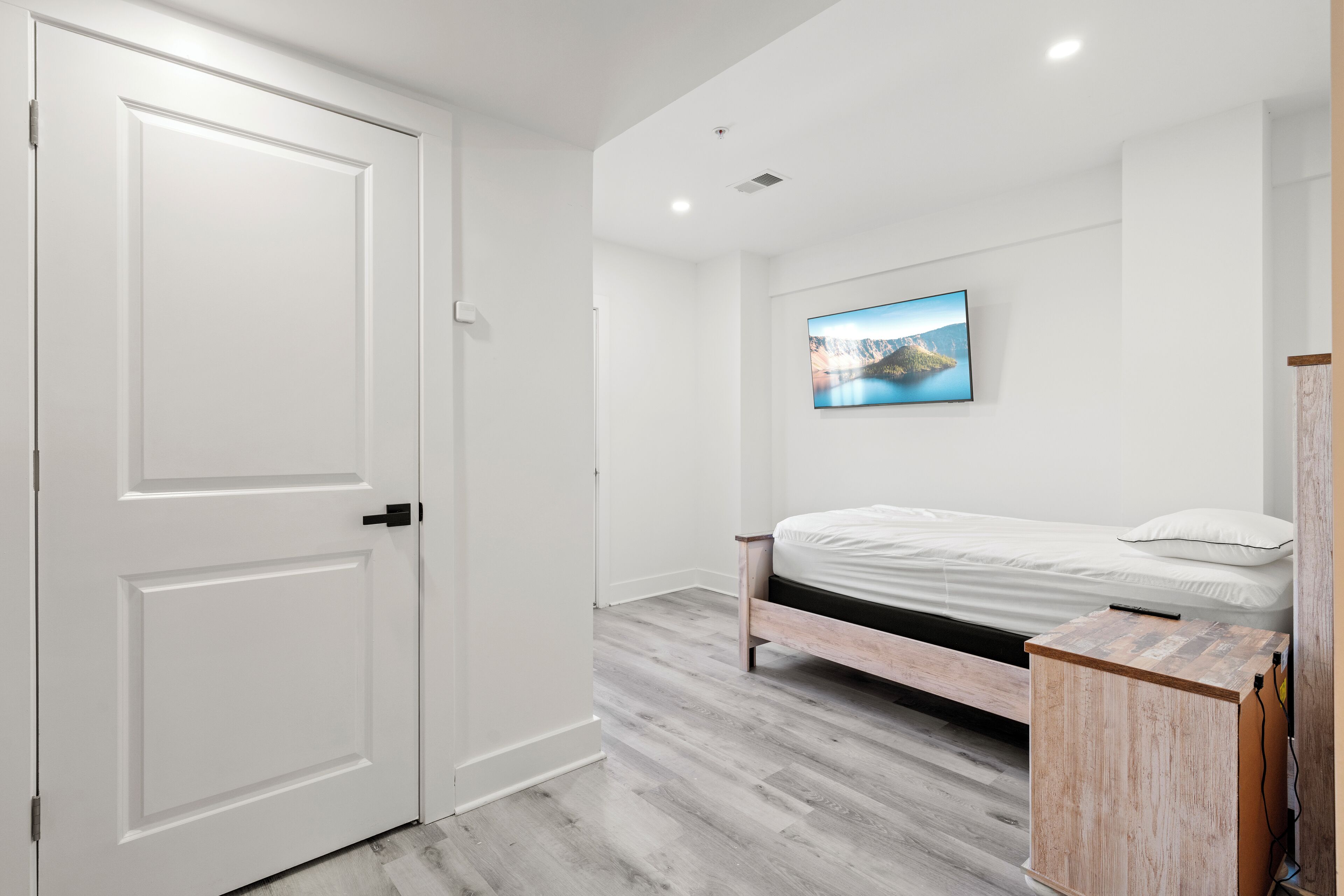 A bedroom with a bed, a white door, a wooden cabinet, and a wall-mounted TV.
