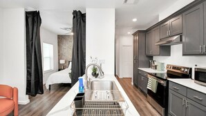 A compact kitchen with a built-in dishwasher, a stove, and a microwave. There is a sink with a faucet and a small countertop area. The kitchen has dark cabinets and a white countertop. - Serenity Studios: Near Medical Center & Museum District (Houston)