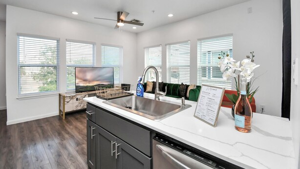 A modern kitchen with a stainless steel sink, a built-in oven, and a dishwasher. There are large windows with blinds, a ceiling fan, and a vase with flowers on the counter. - Serenity Studios: Near Medical Center & Museum District (Houston)