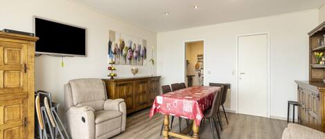A spacious living room with a wooden floor, a dining table with a red tablecloth, a beige armchair, a wooden cabinet, and a flat-screen TV mounted on the wall.