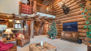 A log cabin interior with a fireplace, leather furniture, a Christmas tree, and a mounted deer head.