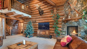 A log cabin interior with a fireplace, a Christmas tree, and a cozy seating area.