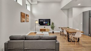 A modern living room with a grey sofa, a wooden dining table with chairs, a flat-screen TV, and artwork on the walls. - 3Bd+65"TV+Crib+8 Min from DT CLE (Cleveland)