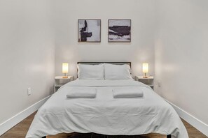 A neatly made bed with white linens and two pillows, flanked by bedside tables with lamps, in a room with plain white walls and a wooden floor. - 3Bd+65"TV+Crib+8 Min from DT CLE (Cleveland)