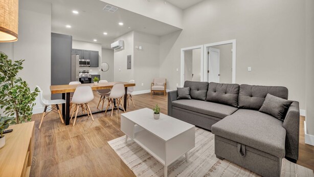 A modern living room with a grey sectional sofa, a white coffee table, and a dining area with a wooden table and white chairs. - 3Bd+65"TV+Crib+8 Min from DT CLE (Cleveland)