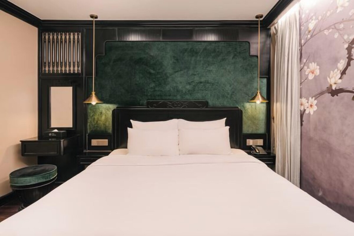 A bedroom with a large bed, two bedside tables, a nightstand, a green upholstered headboard, and a wall with a floral mural.