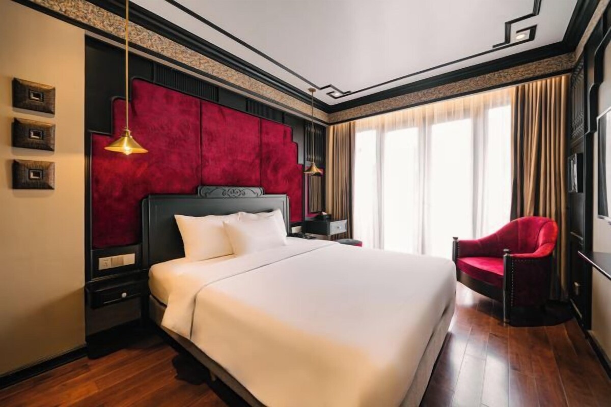A hotel room with a large bed, a red chair, a nightstand, and a window with curtains.