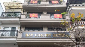 Un hotel llamado "Bay Luxury Hotel" con un diseño arquitectónico clásico, que cuenta con balcones con banderas y una entrada destacada con iluminación decorativa.
