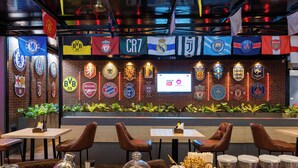 A restaurant with a wall of football club logos, tables with leather chairs, and a digital screen displaying sports content. - The Upper House By Mystical Views (Morjim)