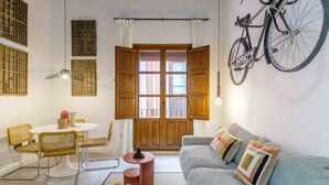 A cozy living room with a sofa, a dining table with chairs, a bicycle mounted on the wall, and a large window with wooden shutters. - Rustic Chic Living4Malaga (Málaga)