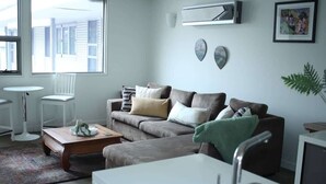Una sala de estar con un sofá gris, una mesa de centro, una silla blanca, una mesita blanca y una zona de cocina con un fregadero.