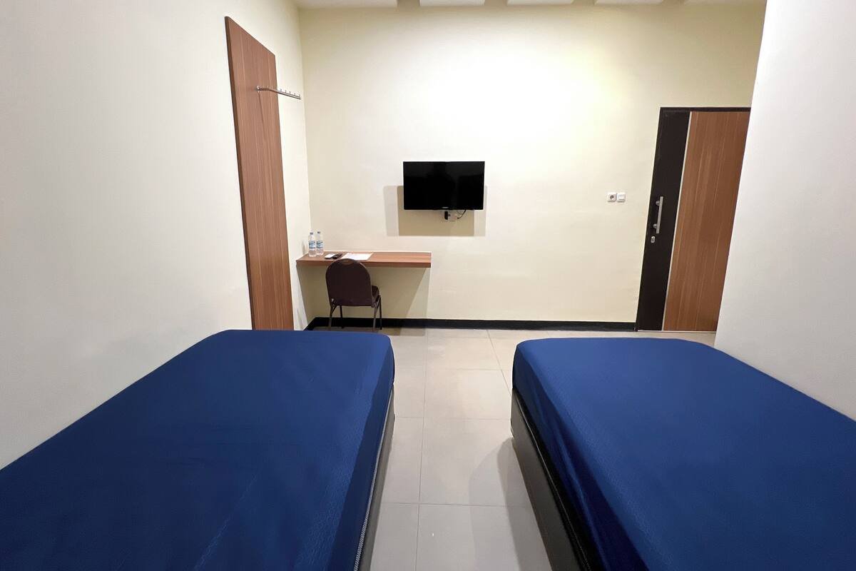 A small, clean room with two single beds, a desk with a chair, a flat-screen TV mounted on the wall, and a wardrobe.