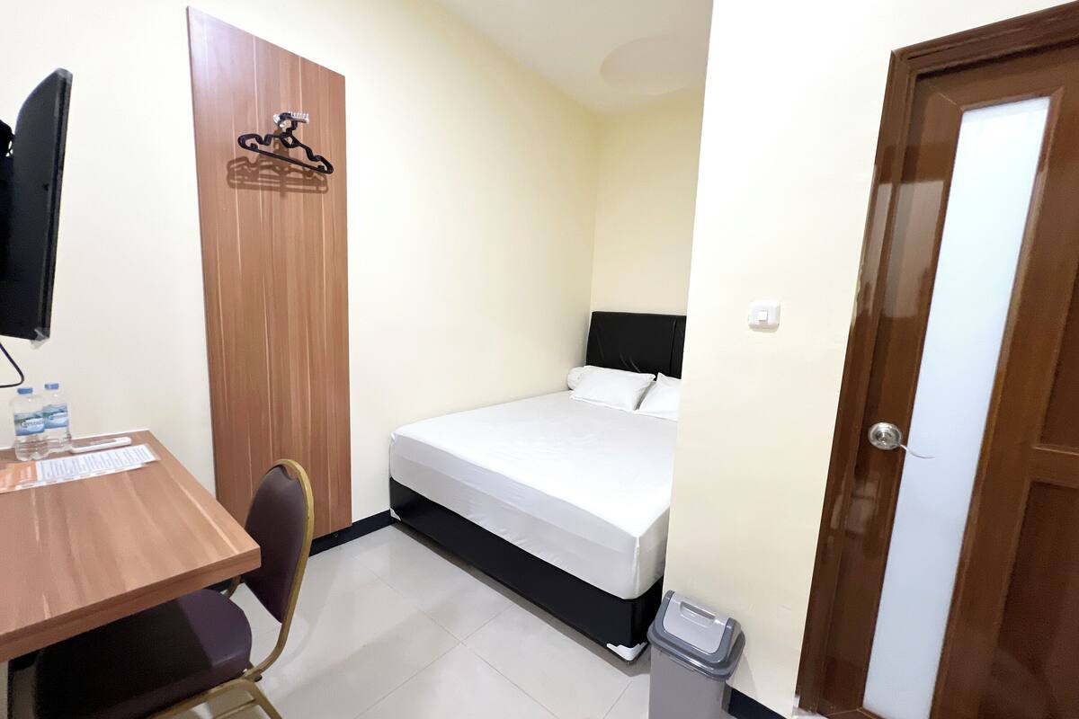 A small hotel room with a bed, a desk, a chair, a TV, a coat rack, and a trash bin.