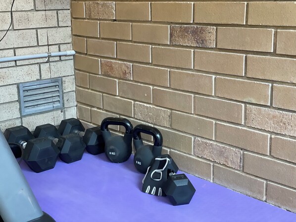 Um conjunto de halteres de 8 kg e kettlebells organizados sobre um tapete roxo, com uma parede de tijolos ao fundo.