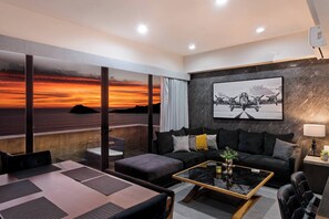 A modern living room with a large window, a black sofa with cushions, a glass coffee table, and a painting on the wall.