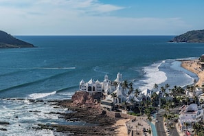 A coastal view with a white building with multiple towers, a beach, and palm trees.