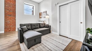 A modern living room with a sectional sofa, a patterned rug, a brick accent wall, a window, and a door.