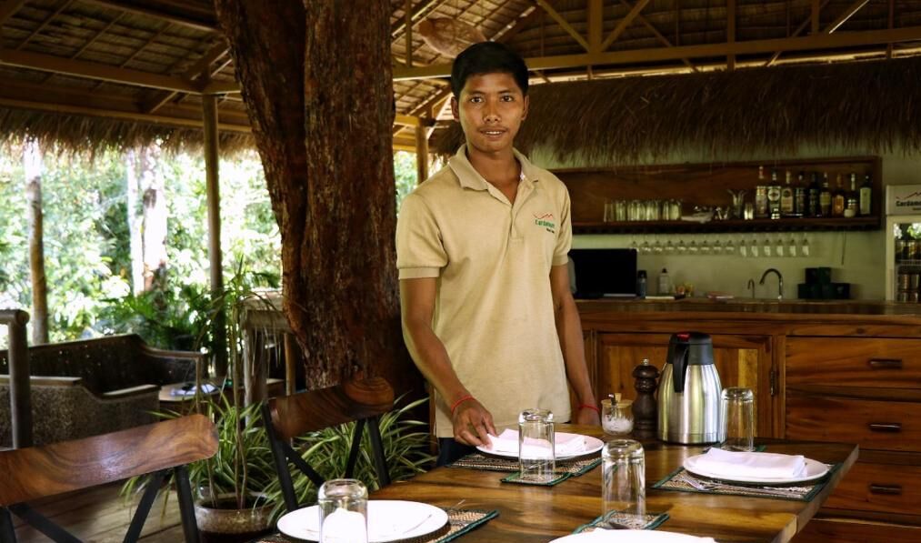 A person standing at a wooden table with plates, glasses, and a coffee pot, in a room with a thatched roof and wooden furniture.